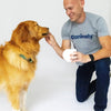Man in a gray 'caninely' shirt feeding a dog from a white bowl on a light background